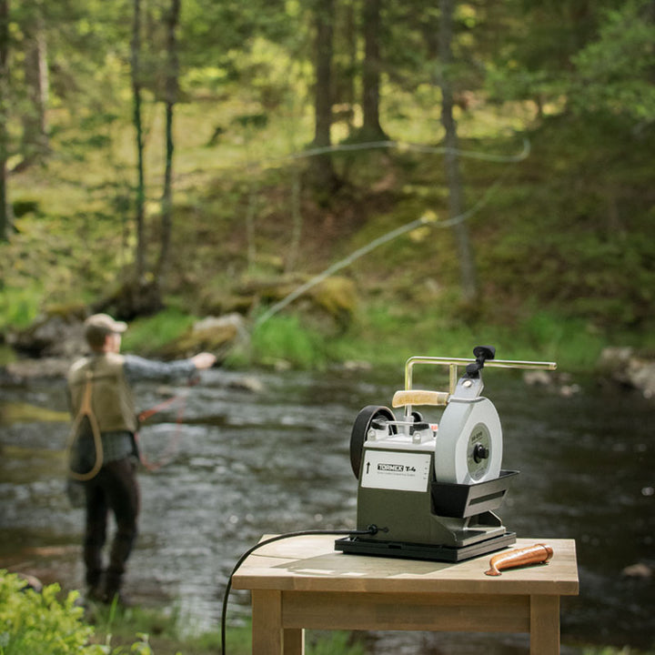 Person fly fishing in a forest stream with a fly fishing reel on a table in the foreground. Sharpening Knives on Tormek T-4 bushcraft machine